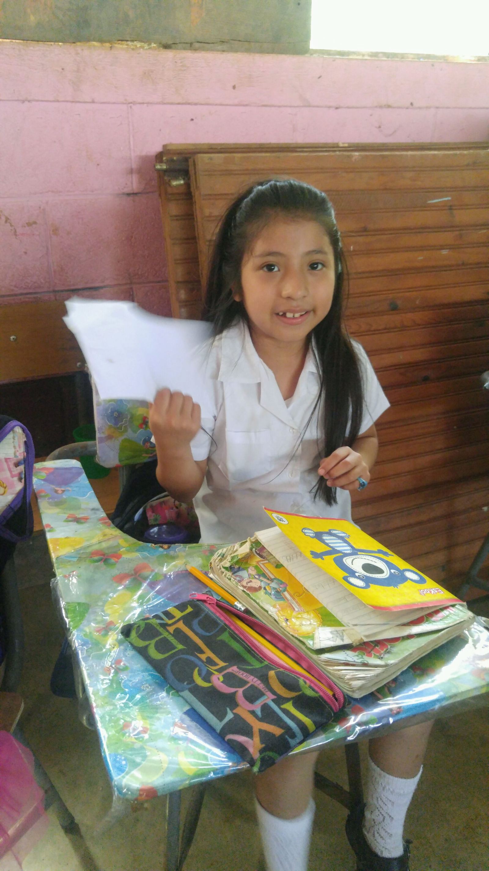 A girl in a white school uniform holds up sheets of paper at her desk beside her pencil case and notebooks