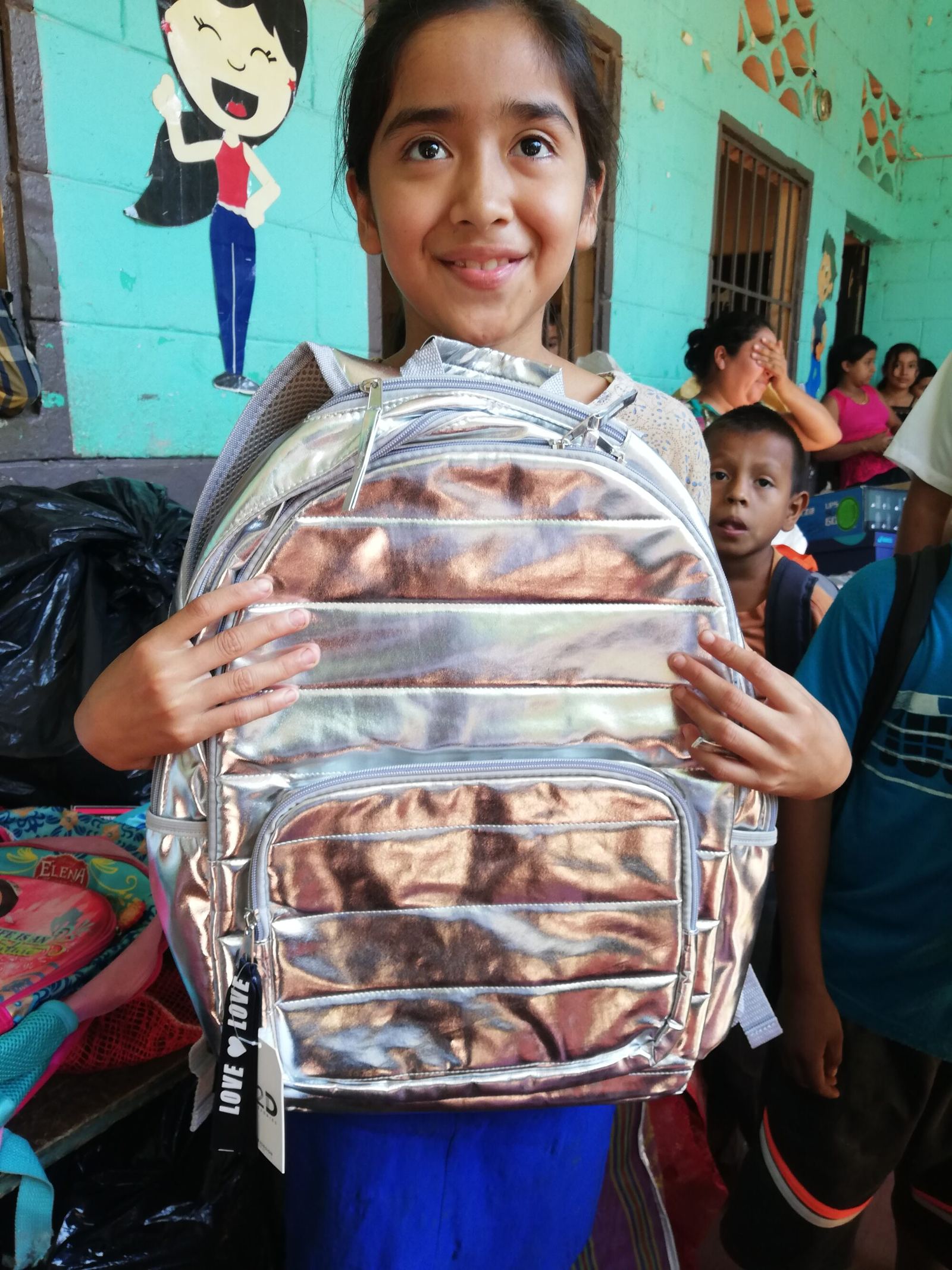 A smiling girl holds up a new silver backpack in front of a school mural
