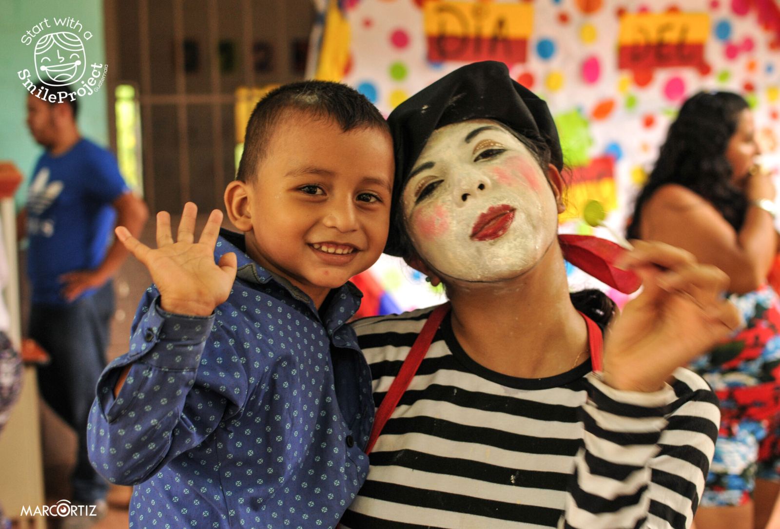 A boy waves at the camera next to a mime in white face paint at a school party