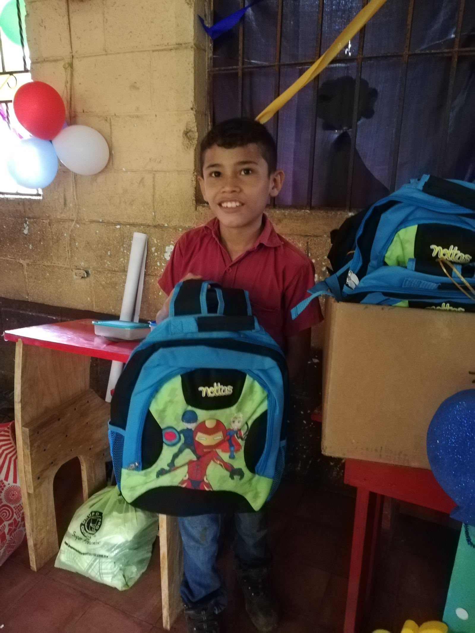 A boy smiles with his new backpack beside a box of more backpacks in a room decorated with balloons