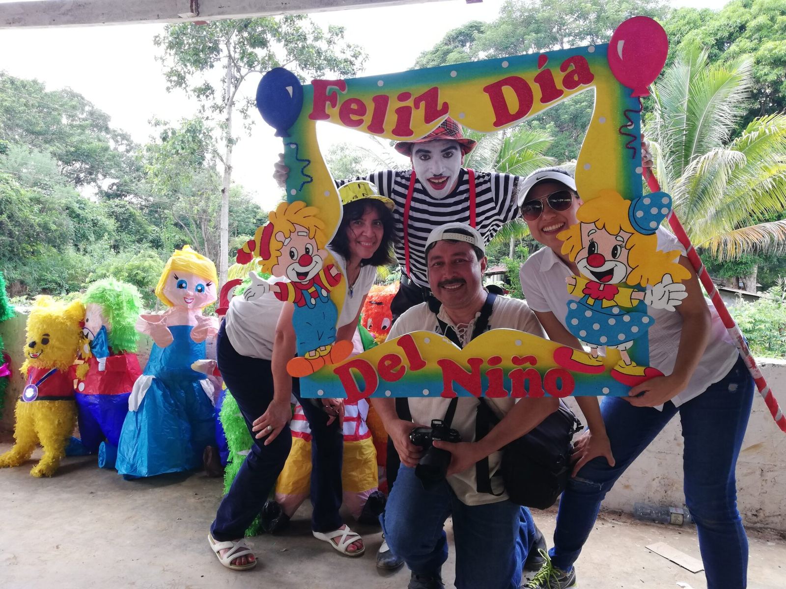 Four adults, one dressed as a mime, pose in a Children’s Day photo frame next to a row of piñatas