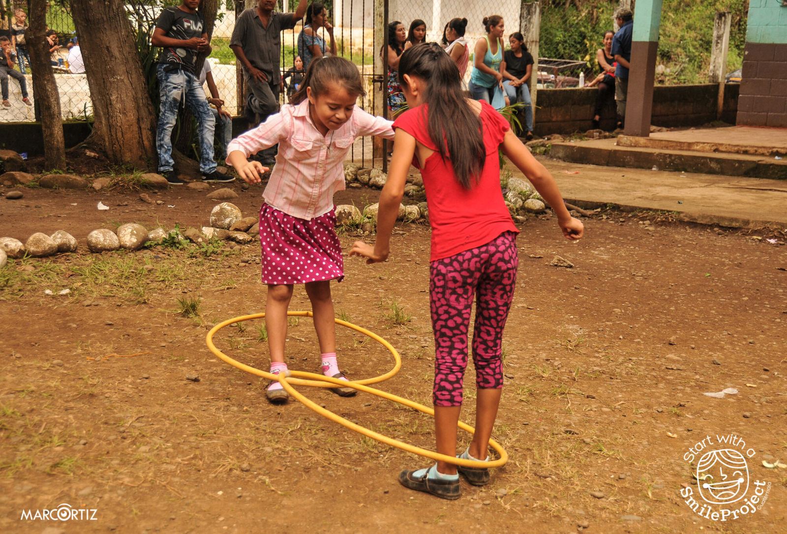 Two girls play with yellow hoops in the dirt schoolyard as teens and adults watch from behind a fence