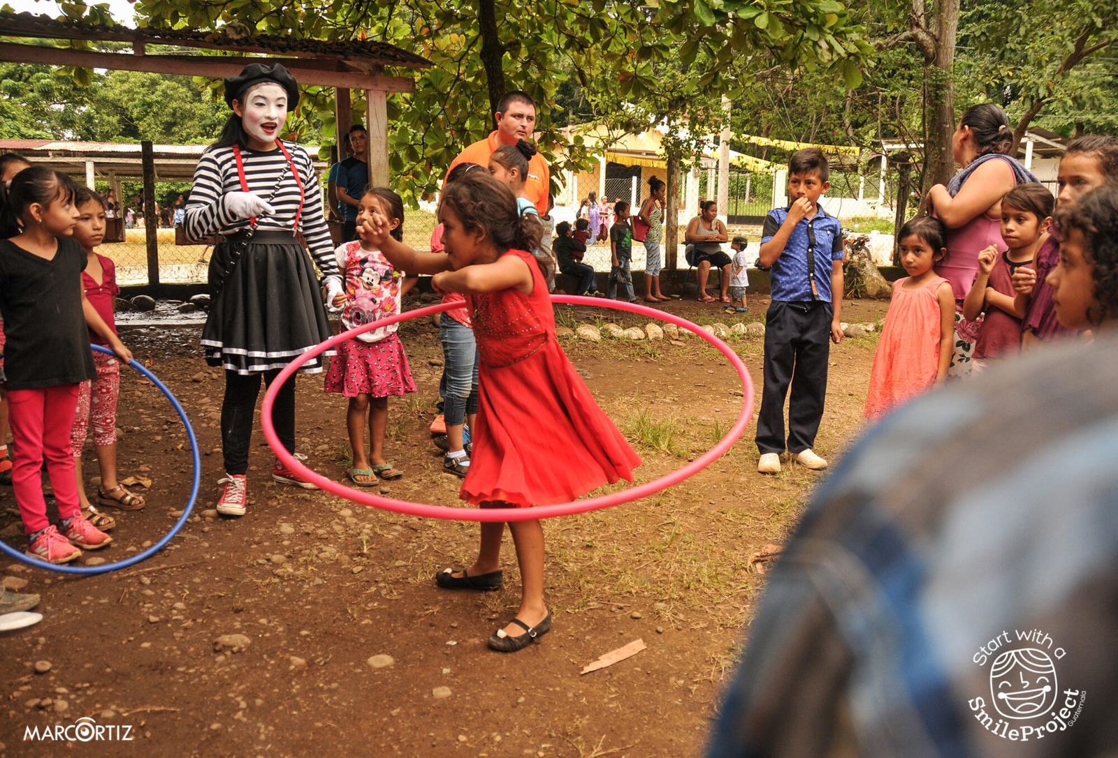 A girl in a red dress spins a hula hoop while a mime and other children cheer her on
