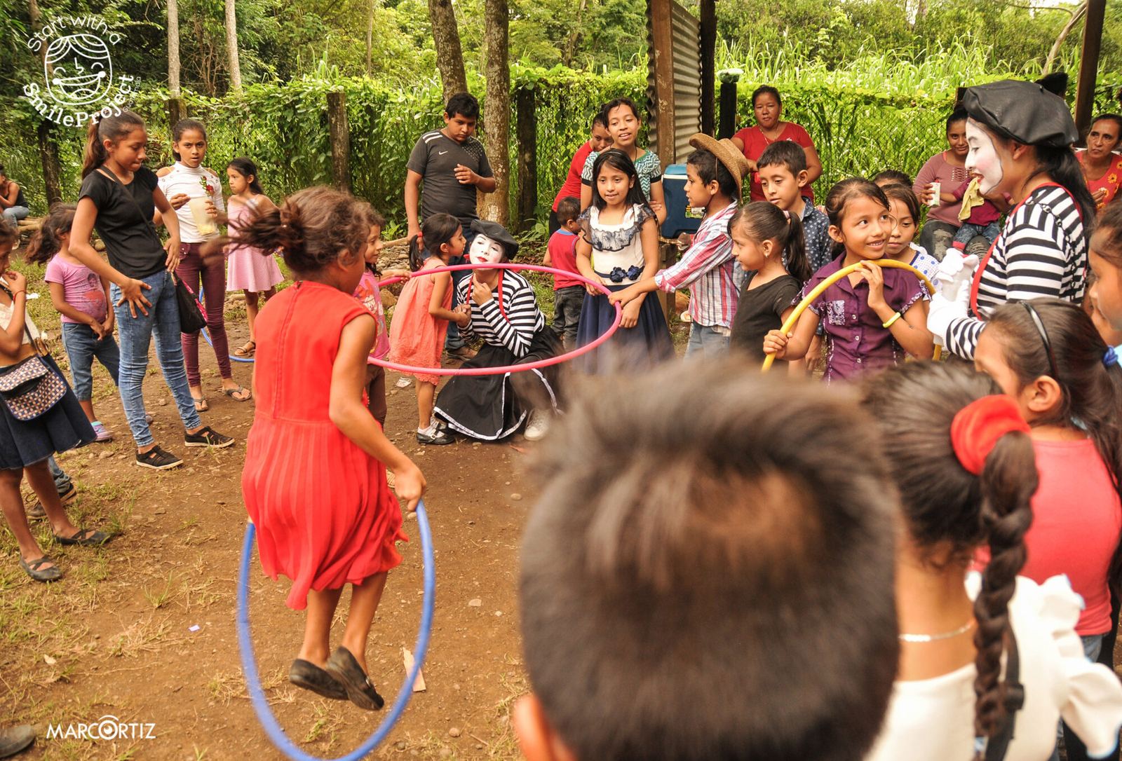 Children in a circle play with colorful hoops alongside two mimes on a dirt field