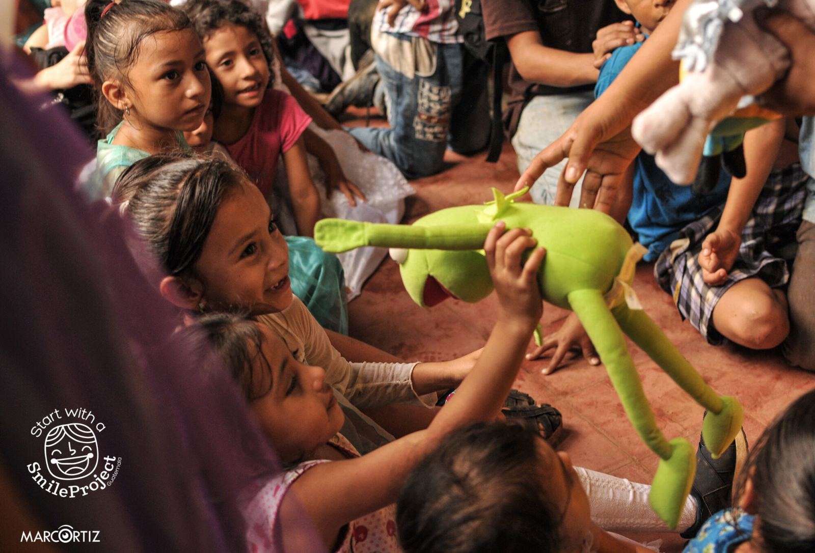 Children sitting on the floor reach for a Kermit the Frog plush toy