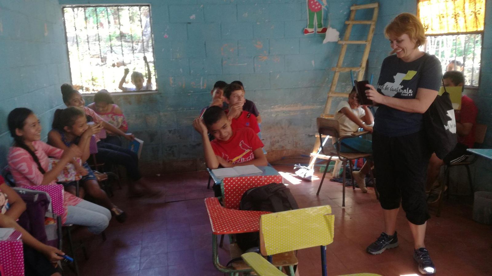 A woman holding a phone smiles at laughing students in a classroom with light-blue walls