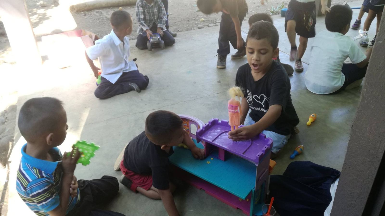 Young children sit on the school corridor floor playing with a dollhouse, a toy truck and bowling pins