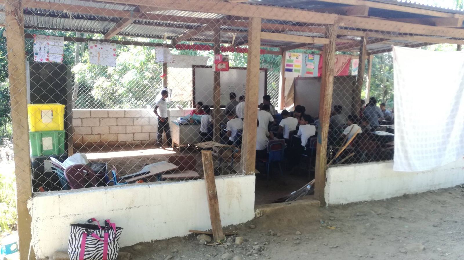 Students attend class in an open-air classroom with a metal roof, wooden posts and chain-link walls