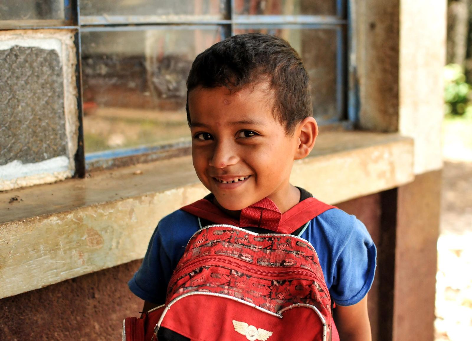 A boy smiles with his red backpack in front of a school window