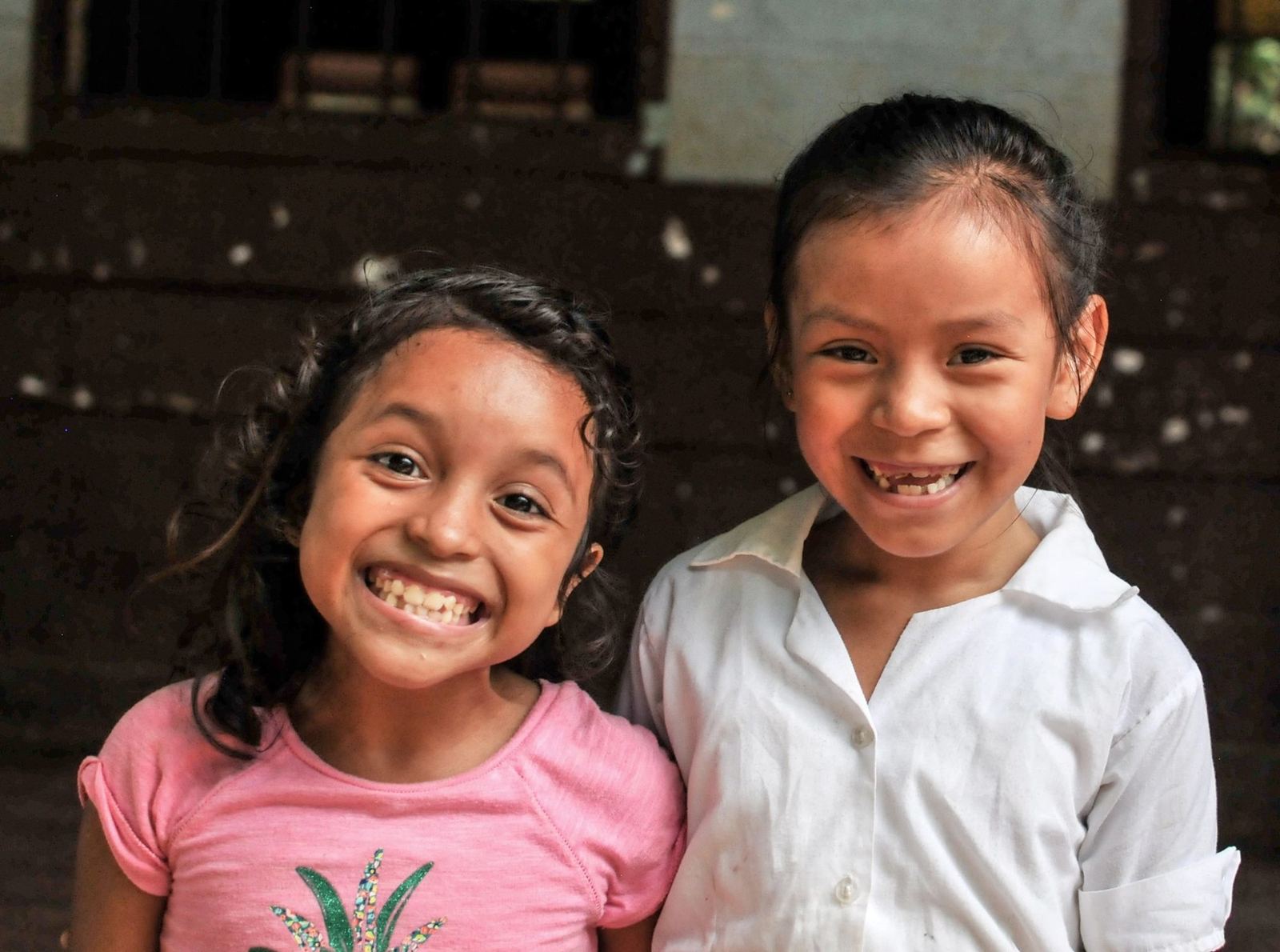 Two girls grin at the camera, one in a pink T-shirt and the other in a white school blouse