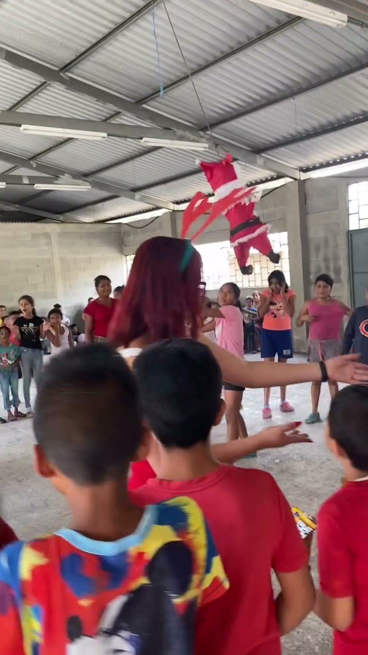 Children in red shirts play a circle game led by a young woman under a Santa Claus piñata