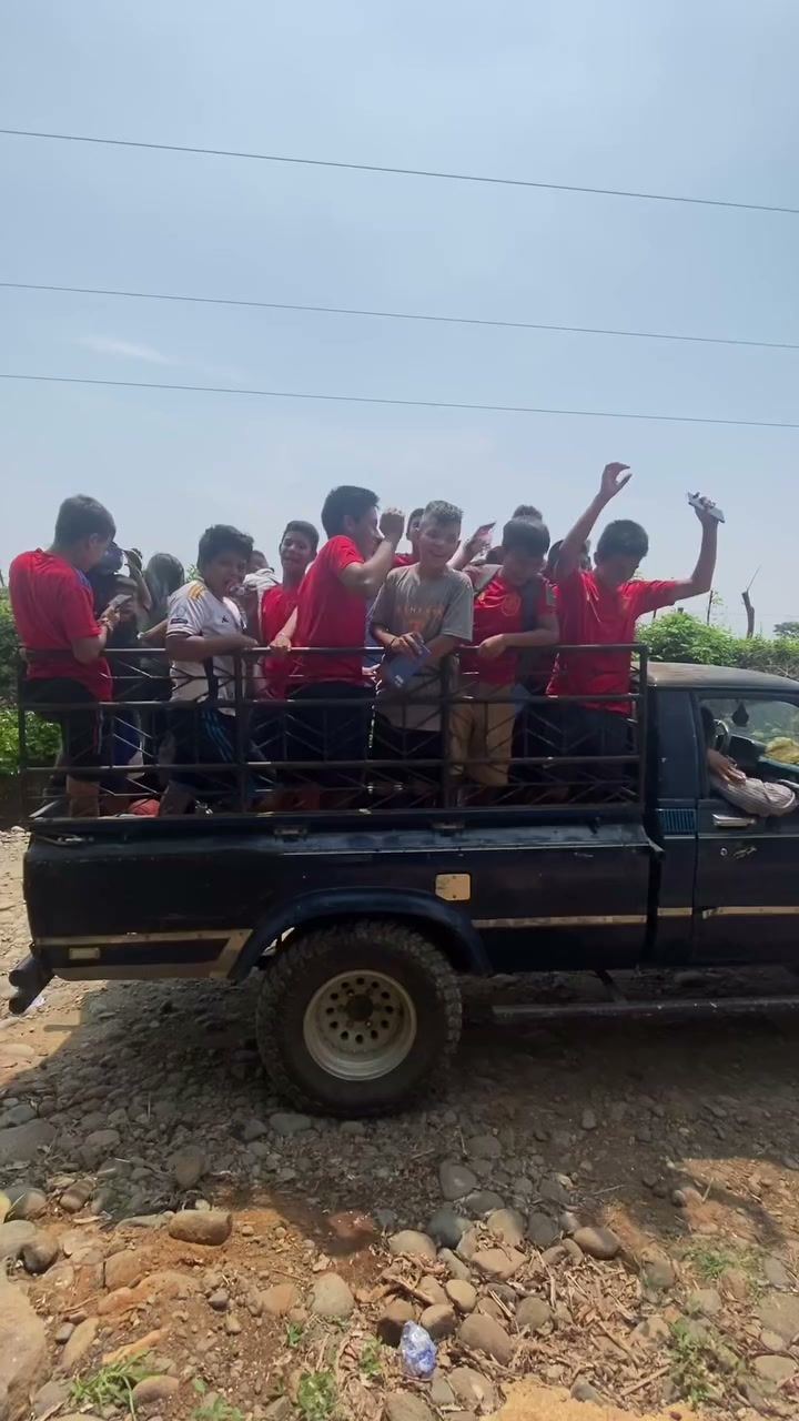 Kids in red soccer jerseys wave from the back of a pickup truck on a dirt road