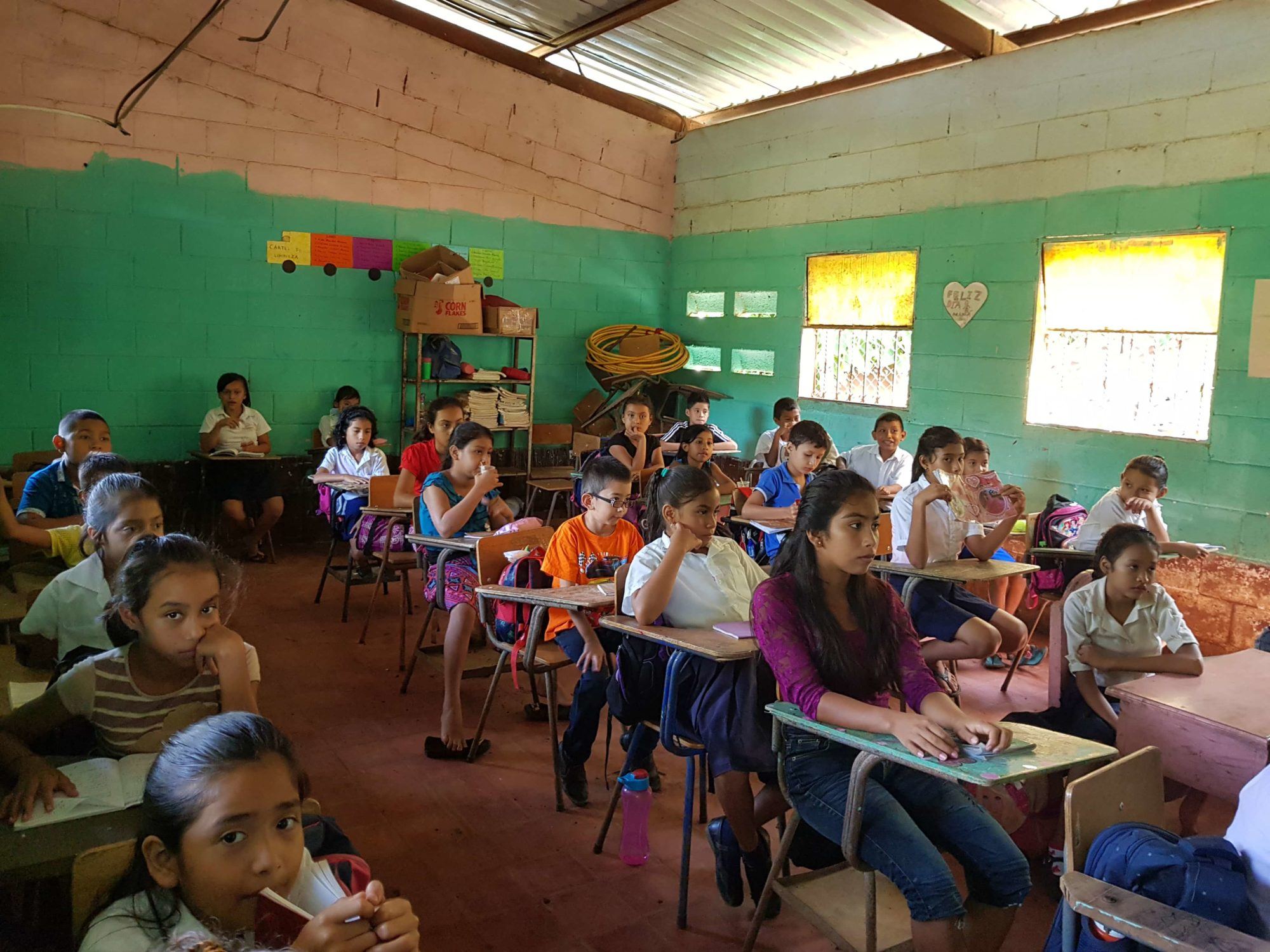 A green-walled classroom packed with students at their desks under a metal roof