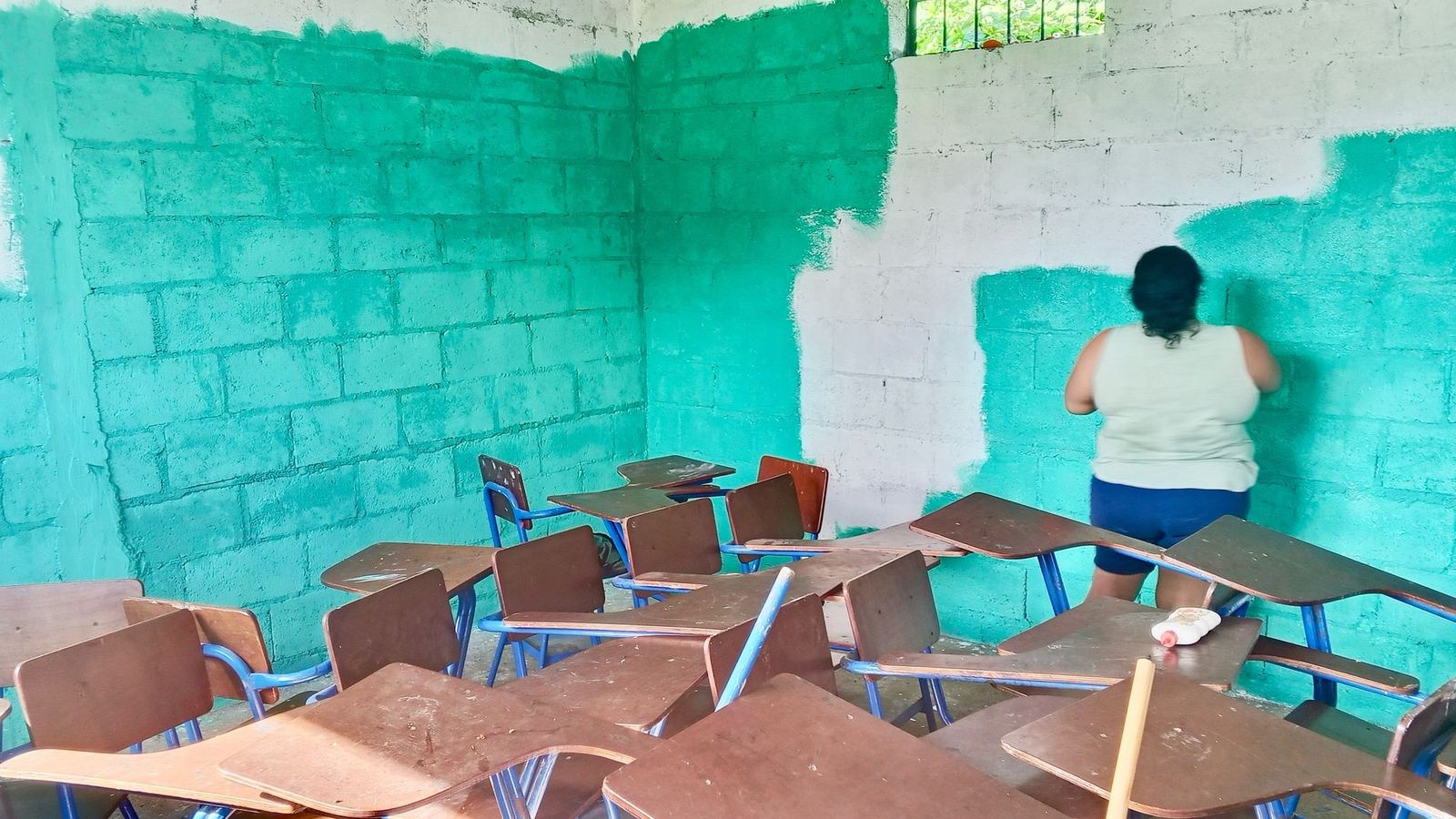 A classroom half painted turquoise in Las Delicias del Jobo, with stacked desks and a woman painting at the back