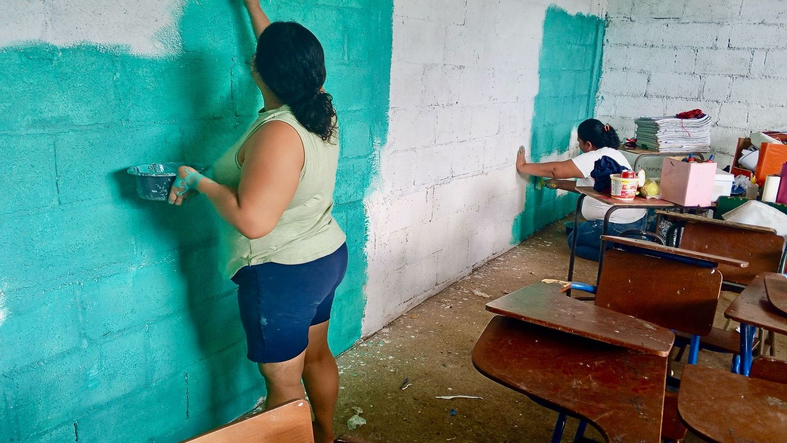Two women paint the block walls of a classroom turquoise in Las Delicias del Jobo, among wooden desks