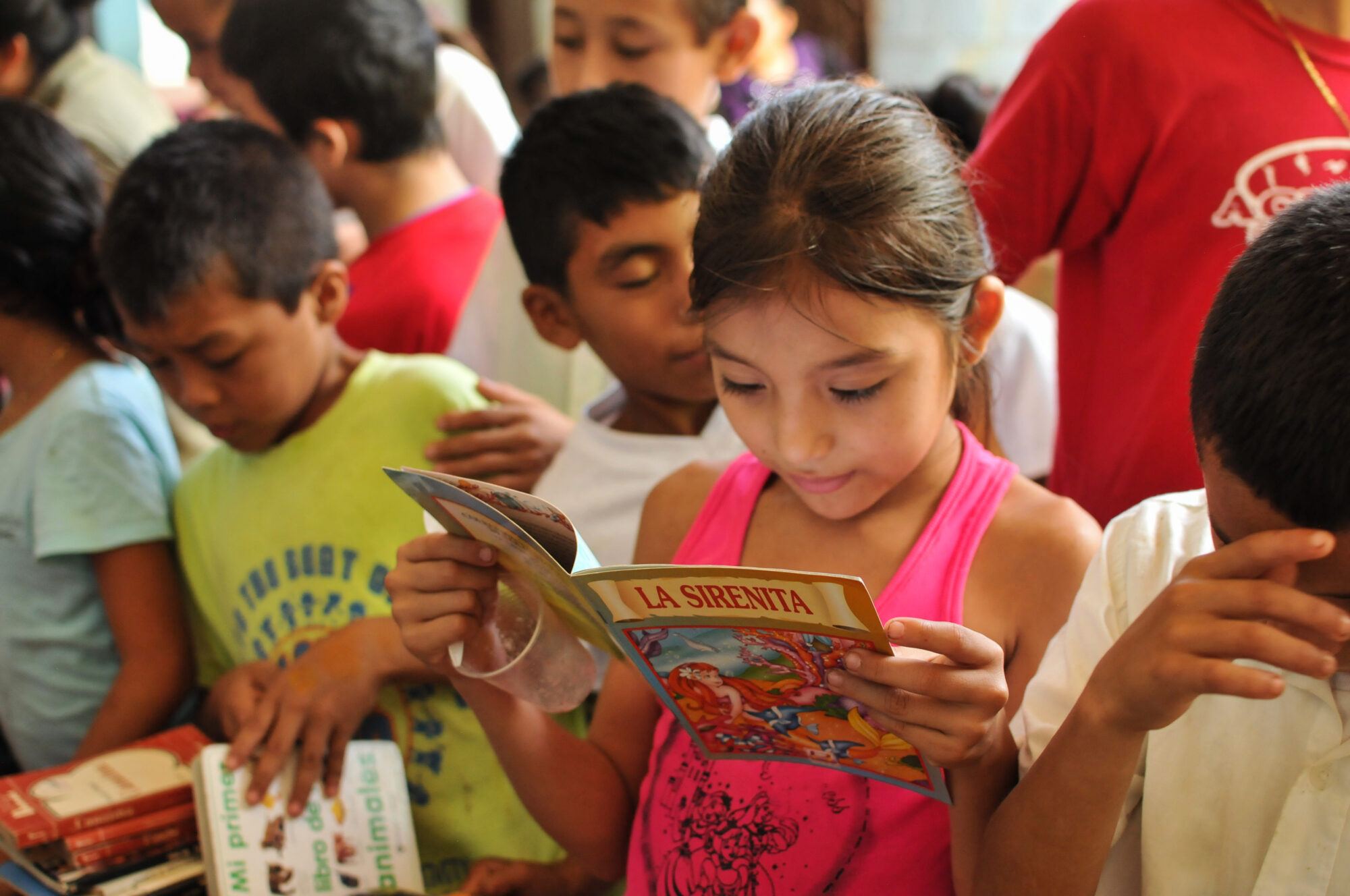 A girl reads “La Sirenita” (The Little Mermaid) among classmates browsing other children's books