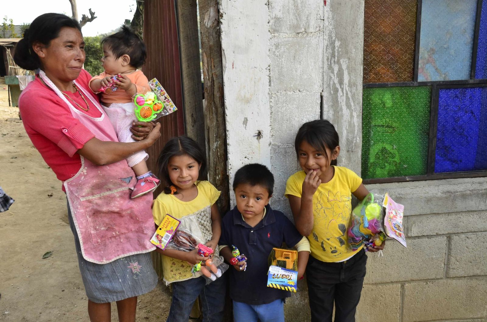 A woman with a baby in her arms stands beside two girls and a boy holding new toys and Christmas candy