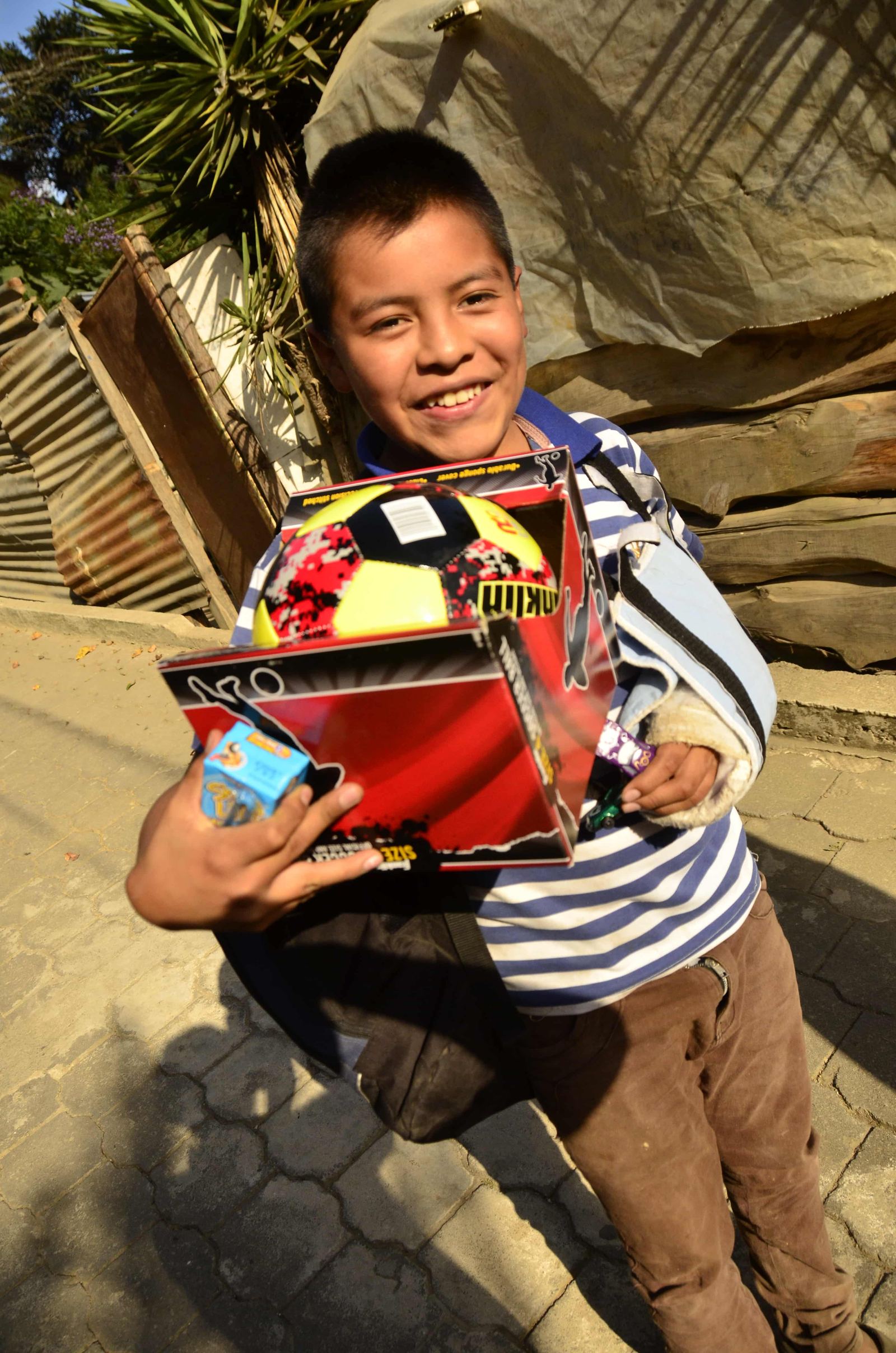A smiling boy holds a boxed soccer ball and other small toys on a paved street