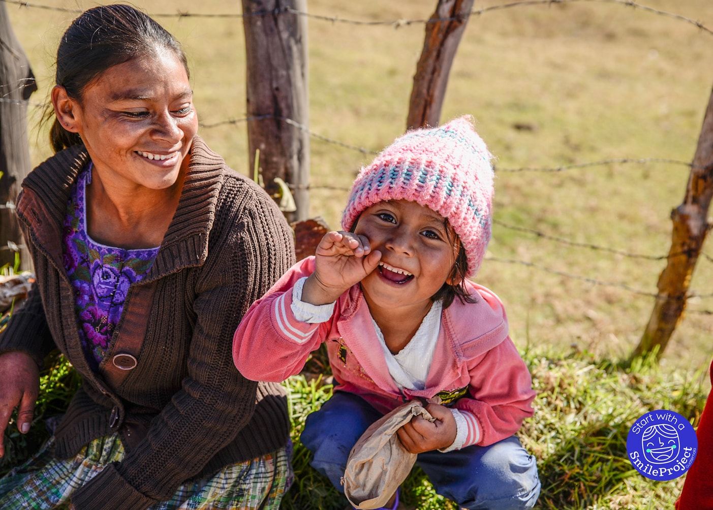 A woman in traditional dress smiles beside a laughing little girl in a pink knit hat, sitting on the grass by a fence