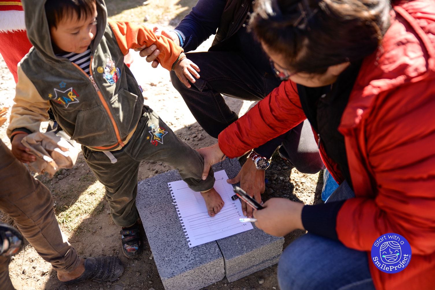 A woman measures a young boy’s bare foot on a sheet of paper to find his shoe size