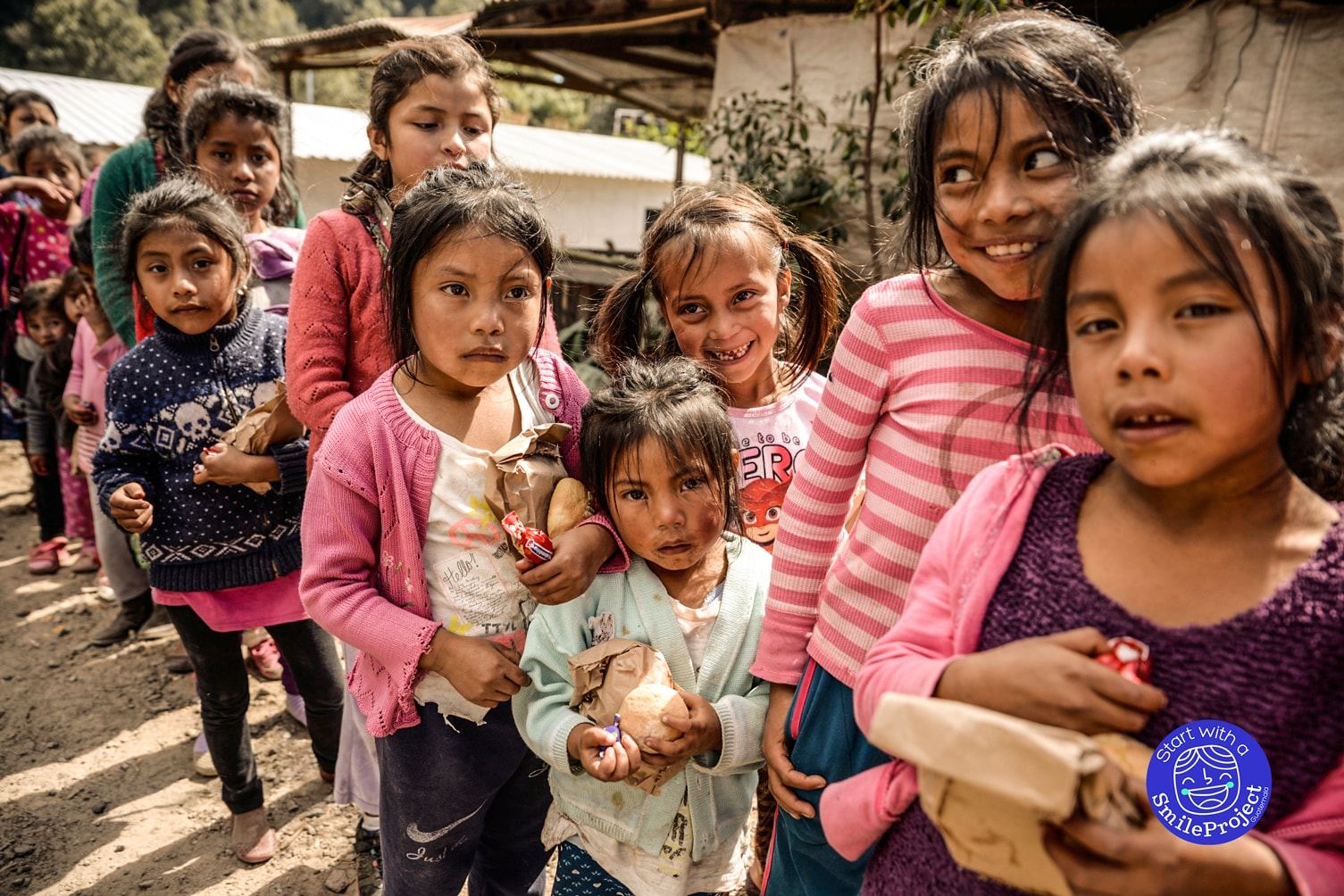 Girls wait in line holding paper bags, bread rolls and candy on a dirt path between houses