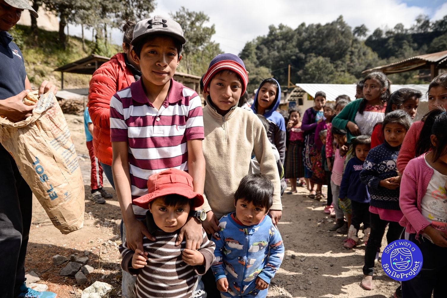 Four boys pose at the front of a long line of children as an adult takes bread out of a sack