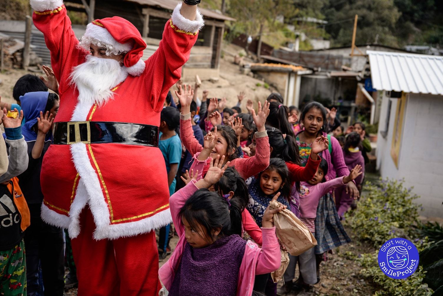 Santa Claus raises his arms and a crowd of children copies him on a dirt path between houses