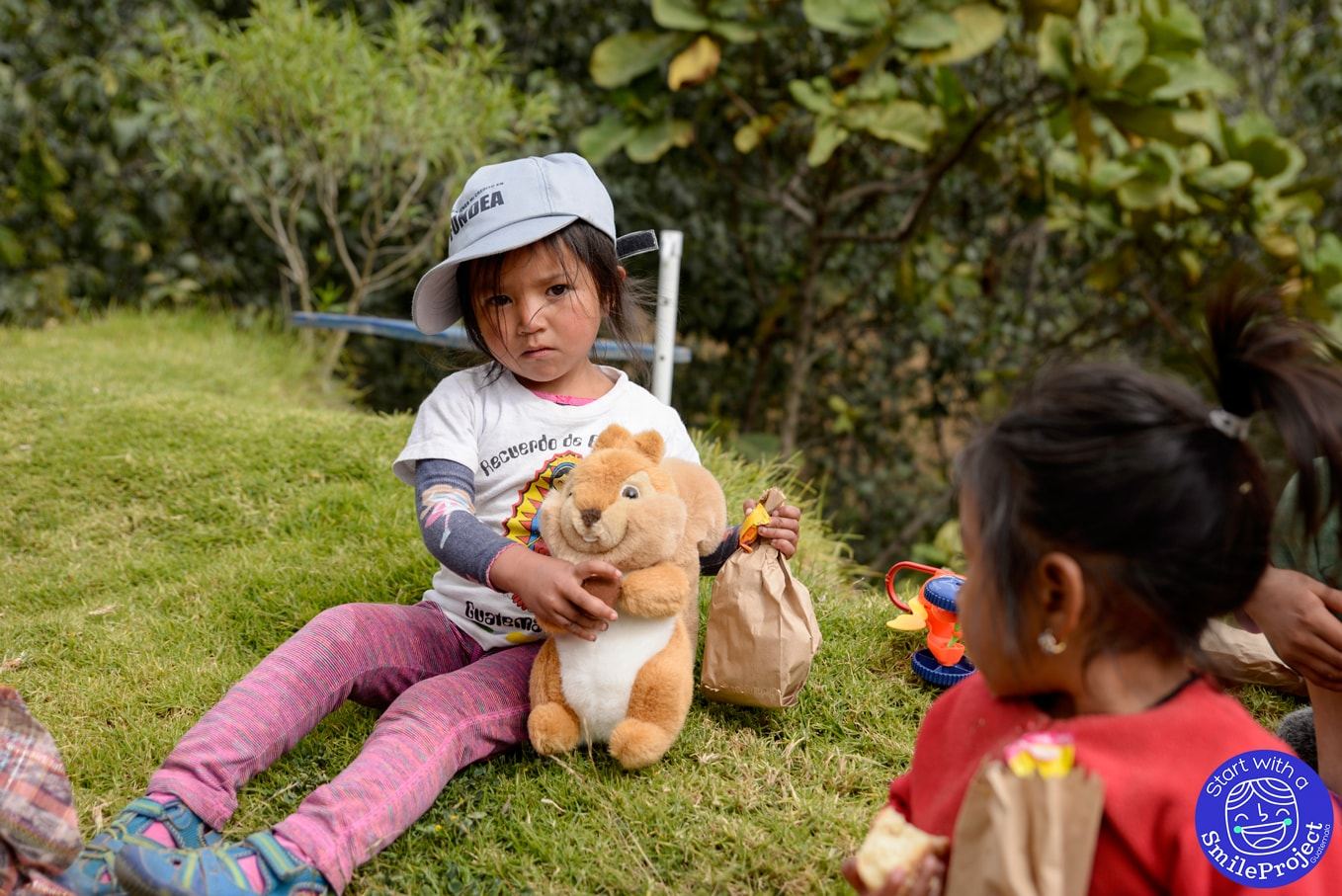 A little girl in a cap sits on the grass hugging a stuffed squirrel and holding a paper bag