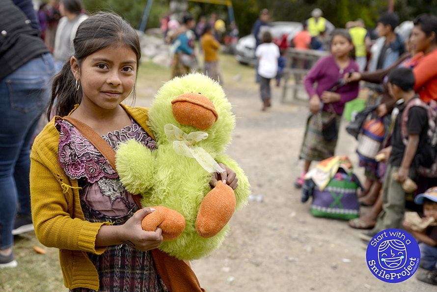 A girl in a mustard sweater hugs a big green stuffed duck, with people gathered on open ground behind her