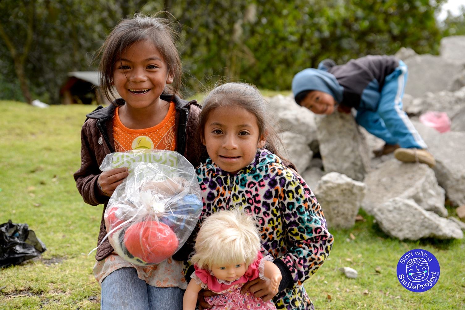 Two smiling girls hold a bag of toys and a doll while a boy climbs on rocks behind them