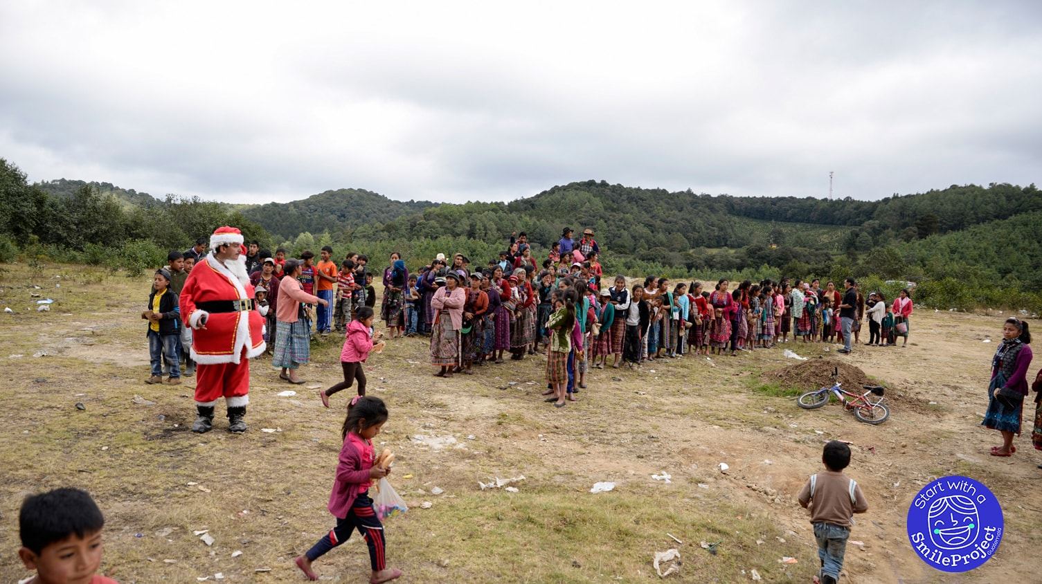 A long line of women and children waits in an open field near Santa Claus, with pine-covered hills behind