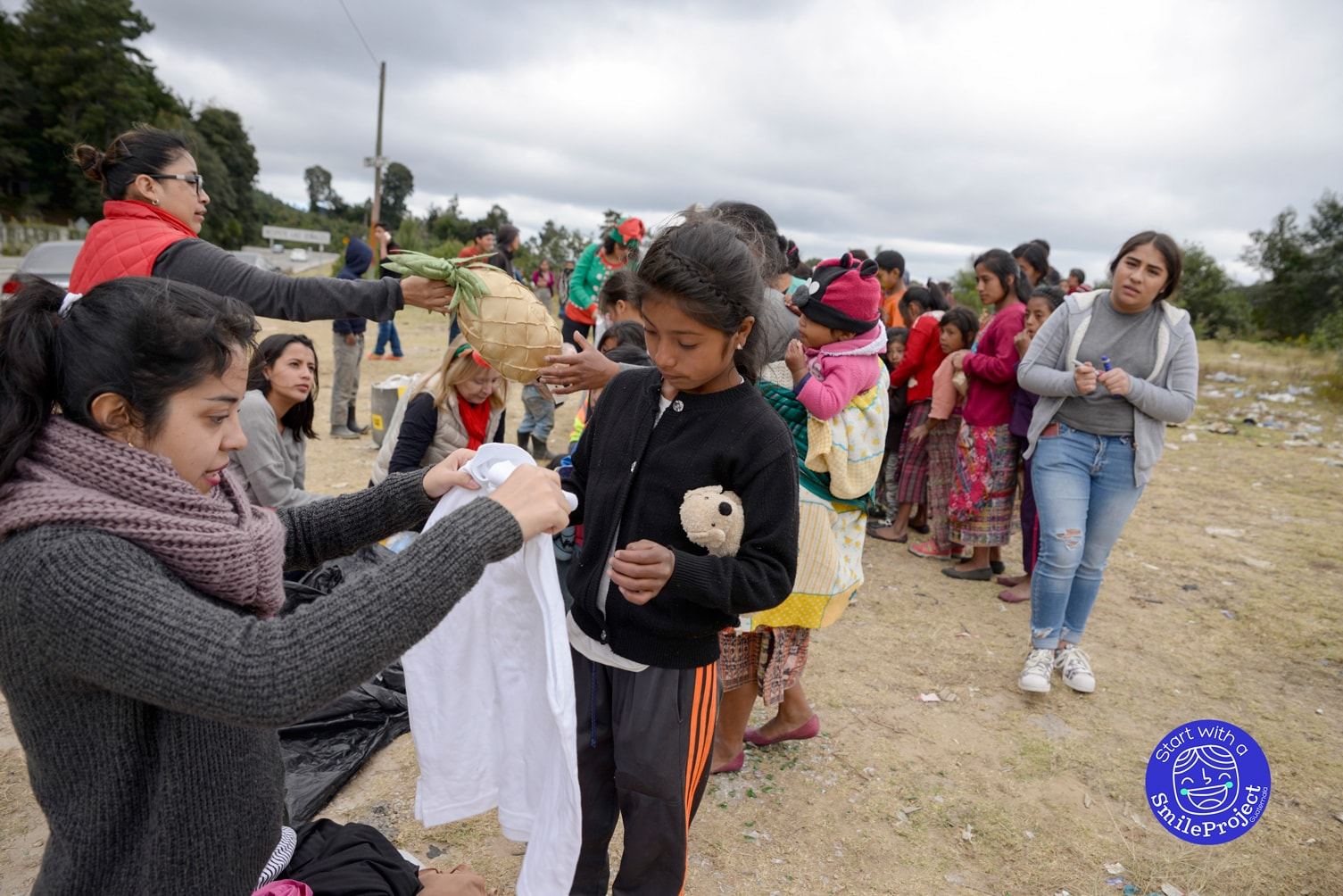 A young woman holds a white shirt up to a girl to check the size as women and children line up by the road