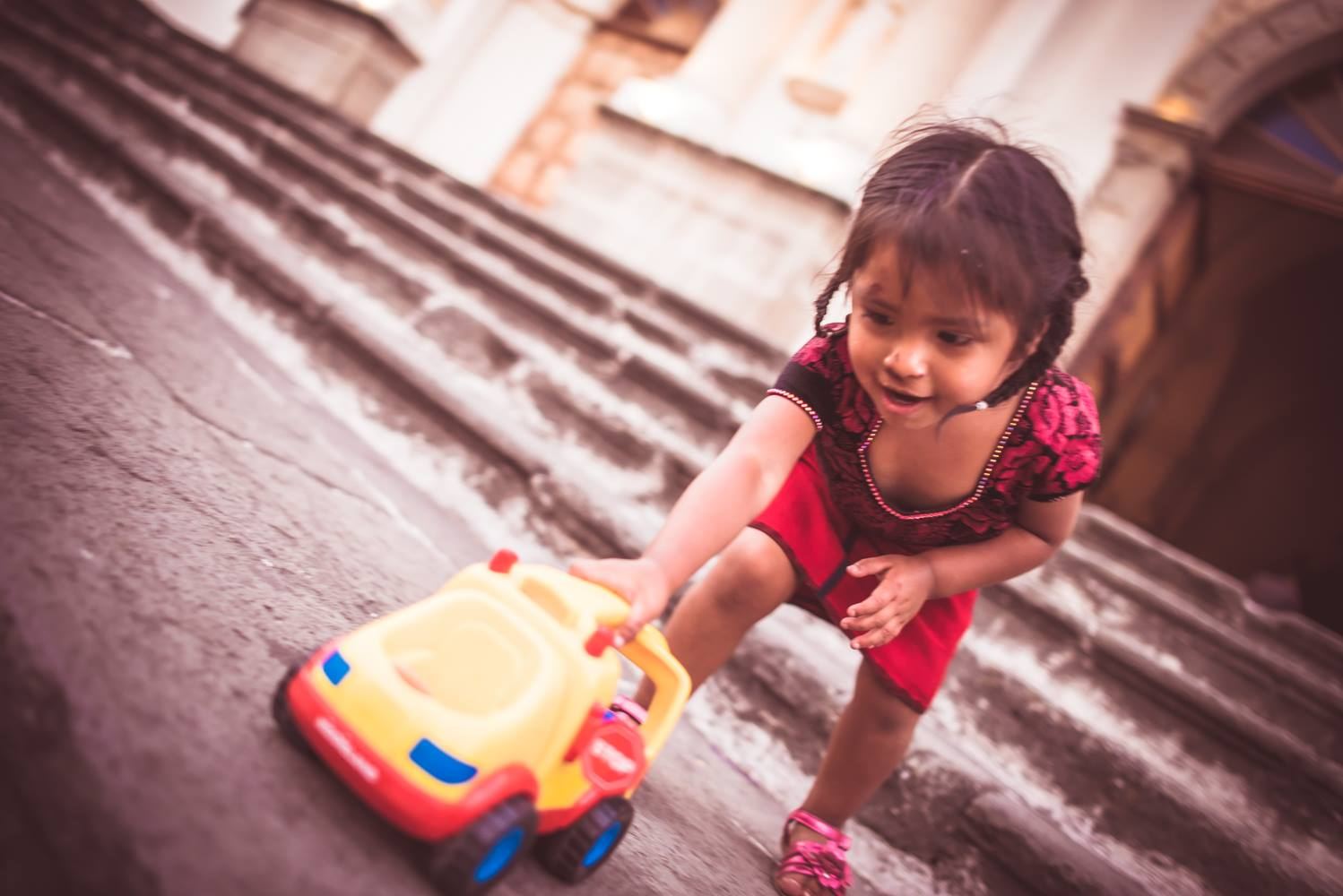 A little girl pushes a yellow and red toy bus in front of the stone steps of a church