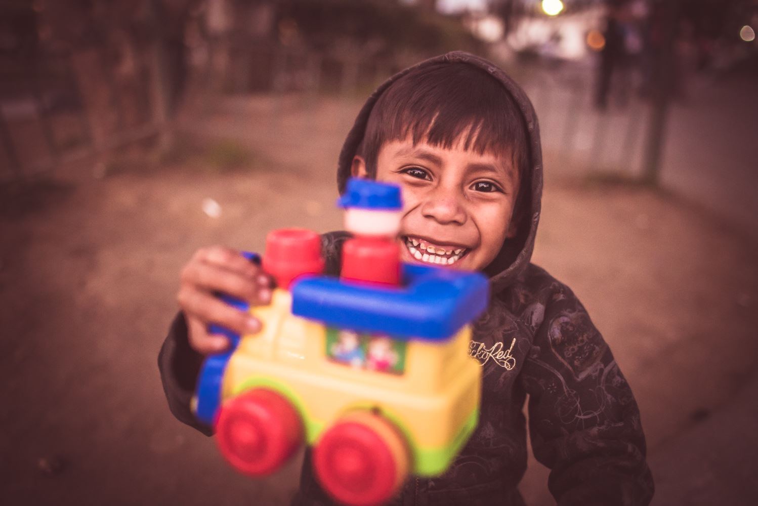 A boy in a hoodie grins as he holds a colorful toy train up to the camera on a dirt street