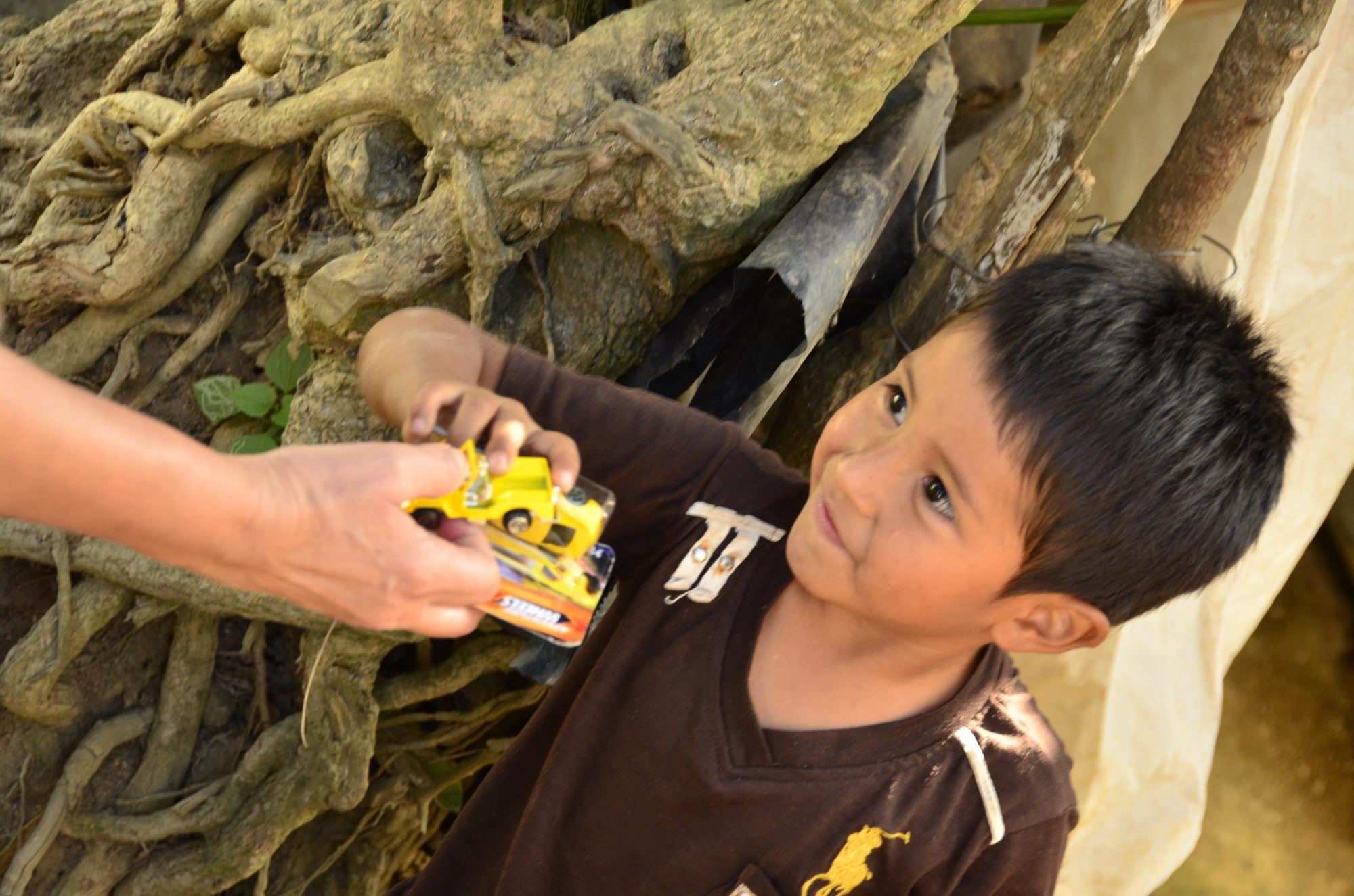 An adult hand passes a small yellow toy car to a boy who looks up attentively