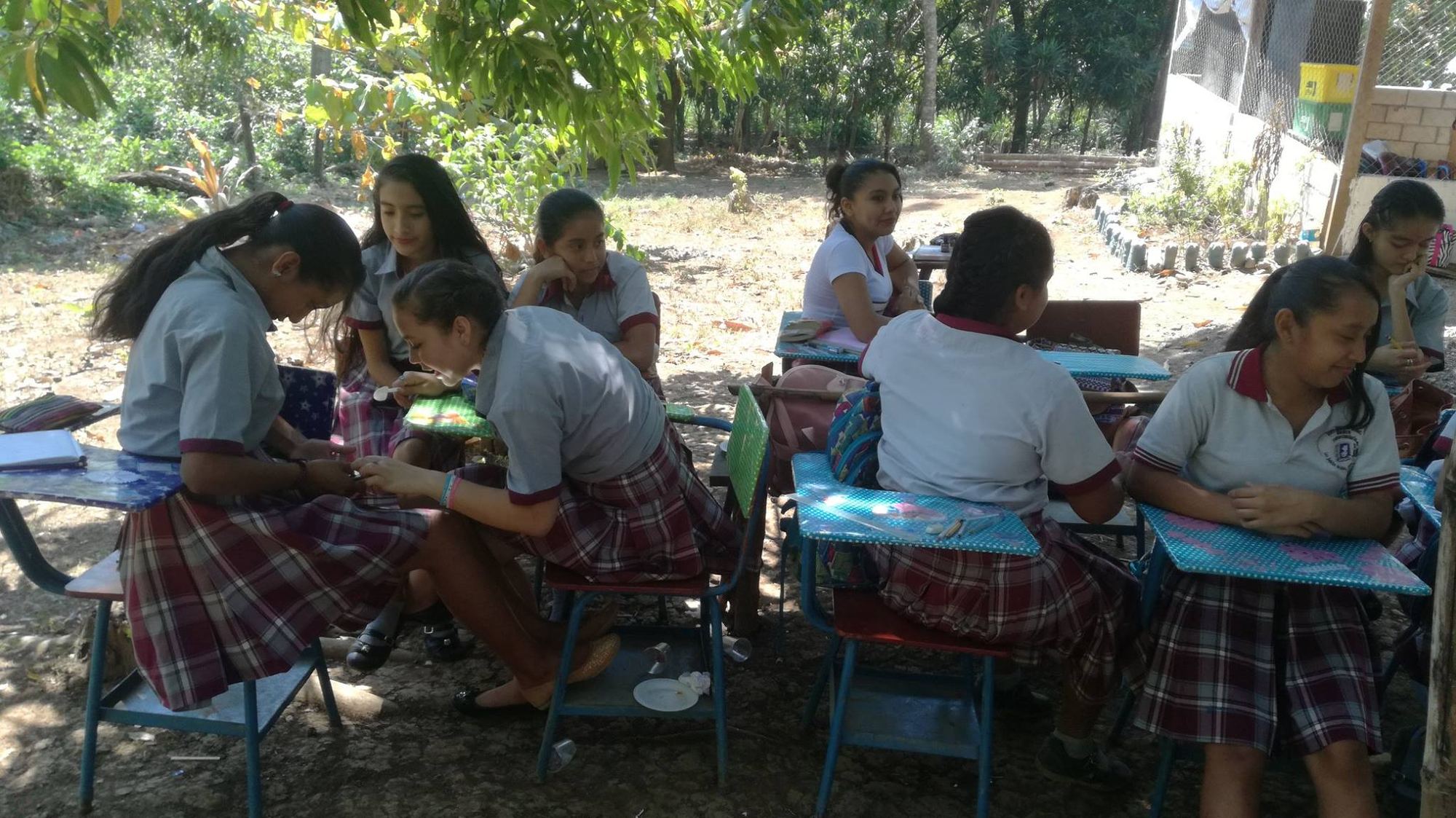 Secondary students in uniform sit at outdoor desks in the shade of trees in Las Delicias del Jobo
