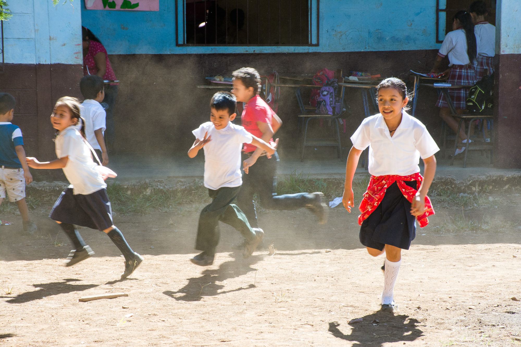 Children in uniform race across the dirt schoolyard in front of light blue classrooms